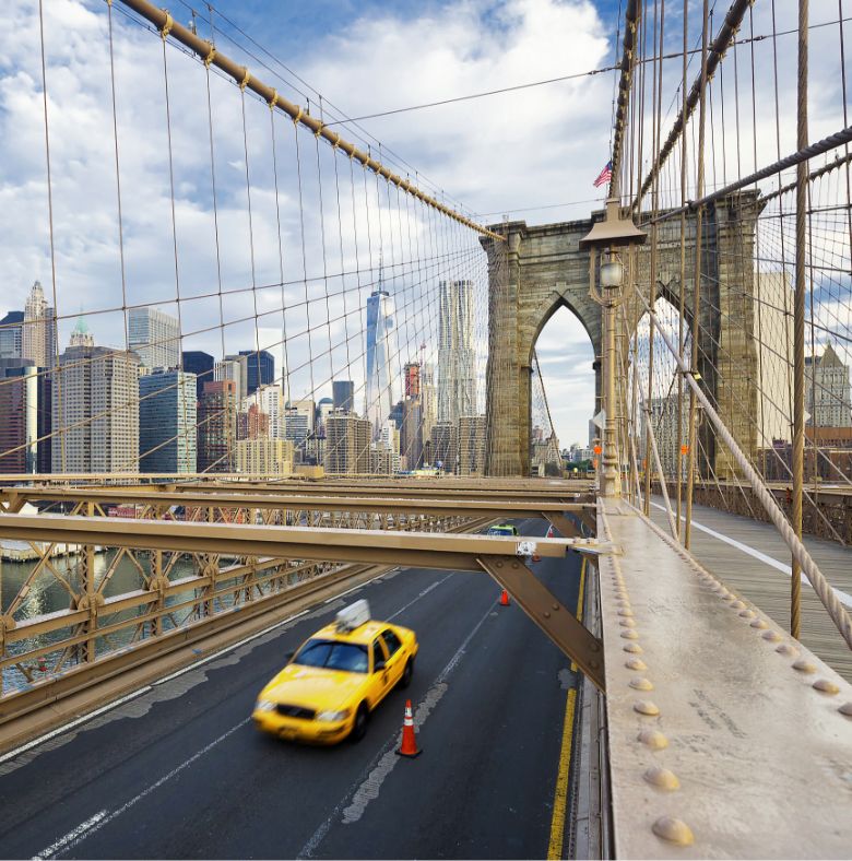 A yellow taxi drives across the Brooklyn Bridge in New York City, with skyscrapers and cloudy blue sky in the background. Suspension cables and bridge structure are visible in the foreground.