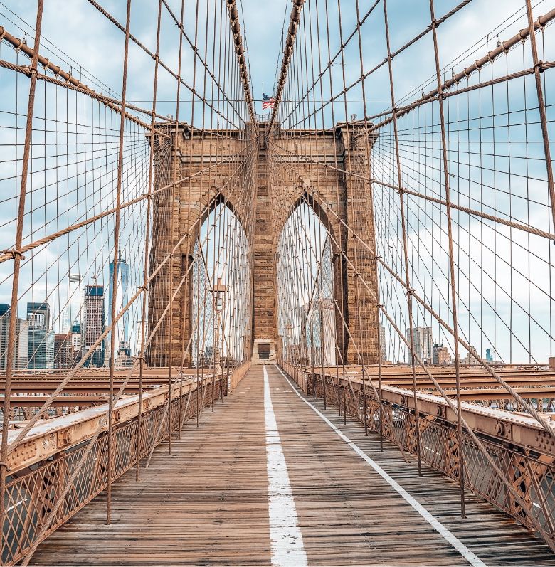 The image shows an empty pedestrian walkway on the Brooklyn Bridge in New York City, with cables and arches visible, and skyscrapers in the background under a partly cloudy sky.