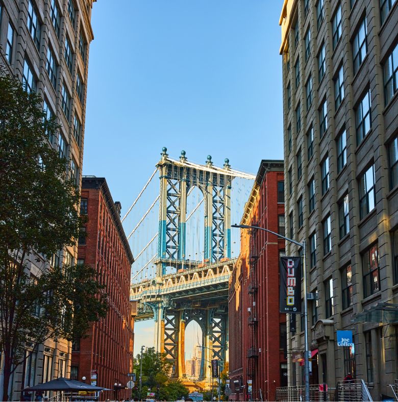 View of the Manhattan Bridge framed by tall brick and stone buildings in Brooklyns DUMBO neighborhood, with trees, street signs, and cars visible on the street below under a clear blue sky.