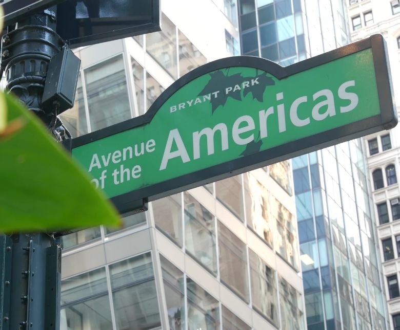 Green street sign reading Avenue of the Americas with Bryant Park above it, surrounded by tall glass buildings and a leafy branch in the foreground.