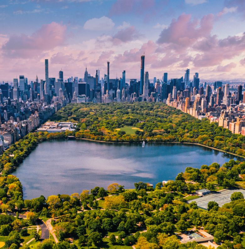 Aerial view of Central Park in New York City, showing a large reservoir surrounded by green trees and skyscrapers under a partly cloudy sky.