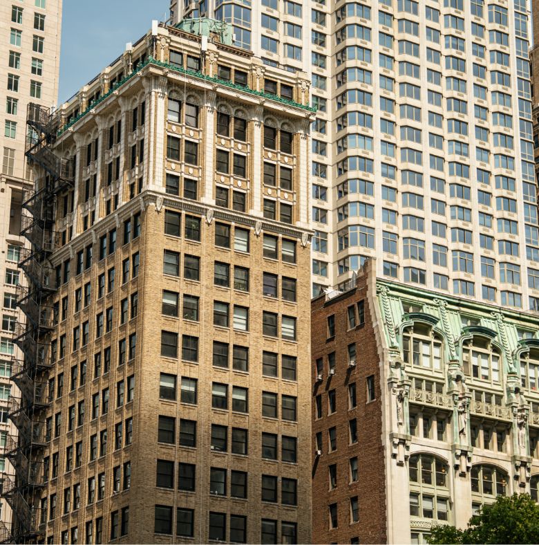 Close-up view of several tall, historic office buildings in a city, featuring ornate architectural details, large windows, and a visible fire escape on one building.