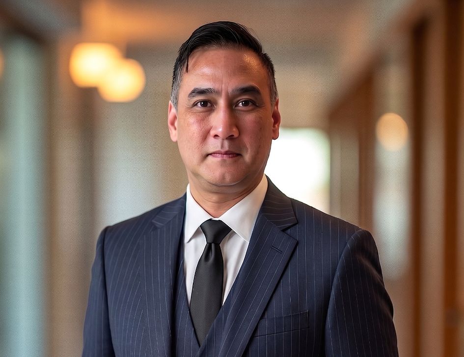 A man in a dark pinstripe suit, white shirt, and black tie stands indoors in a well-lit hallway, looking confidently at the camera.